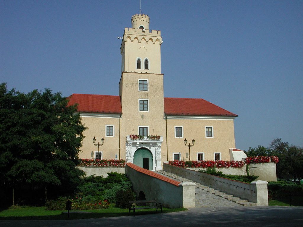 Hrad s vežou a červenými strechami, obklopený kvetmi a stromami, pod jasnou modrou oblohou.