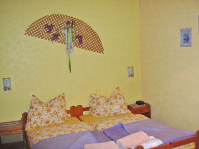 Útulná spálňa so žltými stenami, manželskou posteľou so vzorovanými vankúšmi a dekoratívnym nástenným ventilátorom.