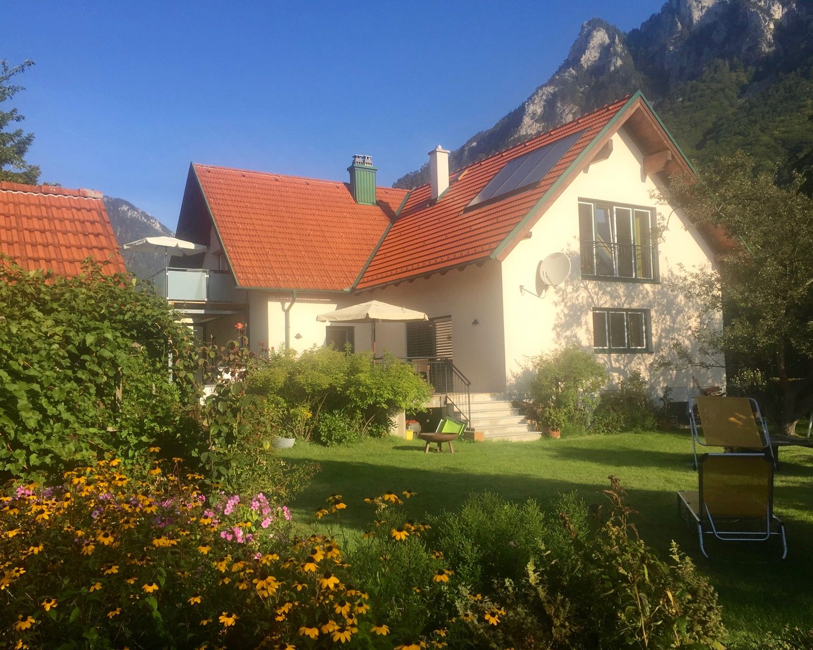 Dom s červenou strechou, obklopený záhradou s kvetmi a ležadlami, na pozadí hôr.