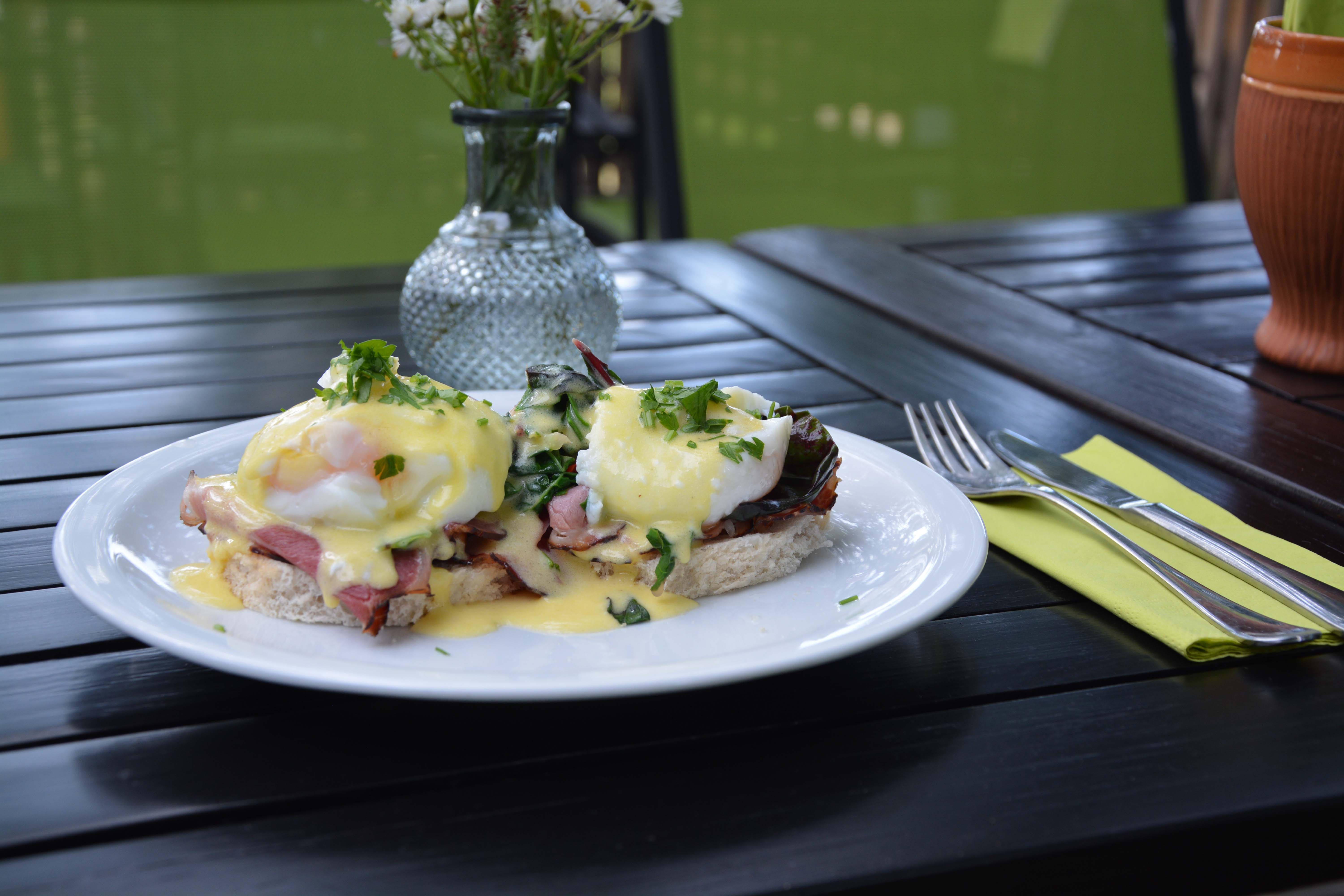 Na drevenom stole tanier s vajíčkami Benedikt, vedľa príbor a váza s kvetmi.