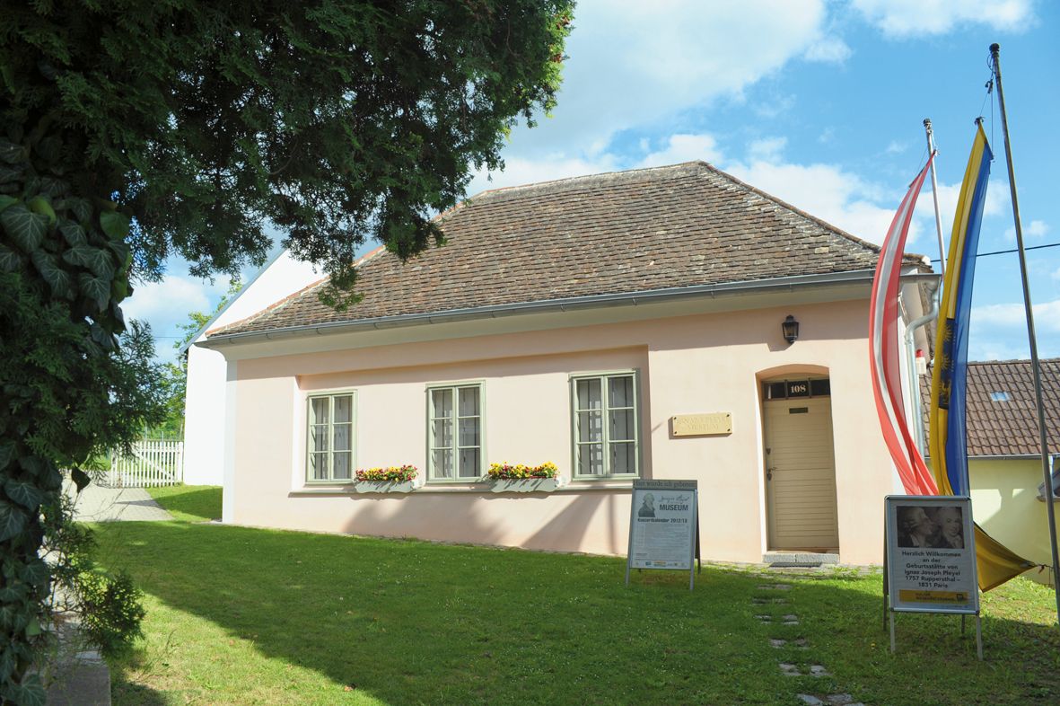Malá, ružovo natretá budova so šindľovou strechou, obklopená stromami a vlajkami, s nápismi pred vchodom.