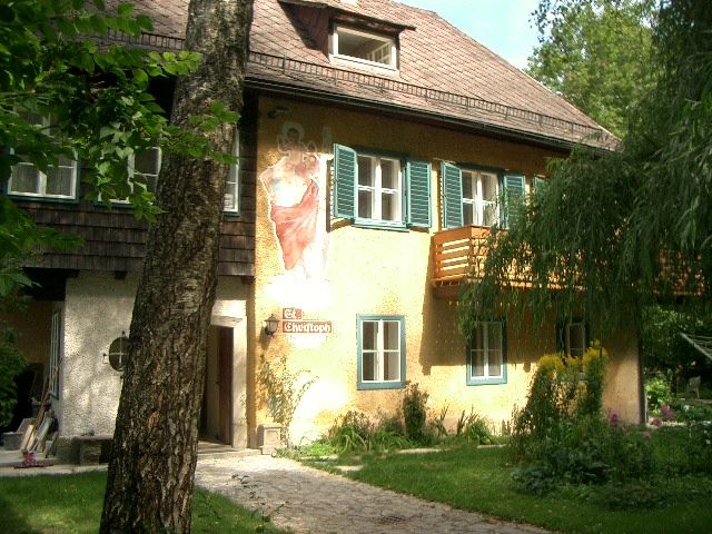 Dvojposchodový dom so žltou fasádou, zelenými okenicami a nástennou maľbou. V popredí stojí strom.