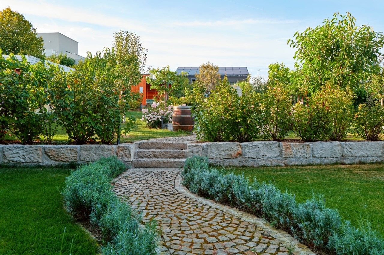 Udržiavaná záhrada s kamennou cestičkou, kríkmi a stromami, v pozadí budova so solárnymi panelmi.