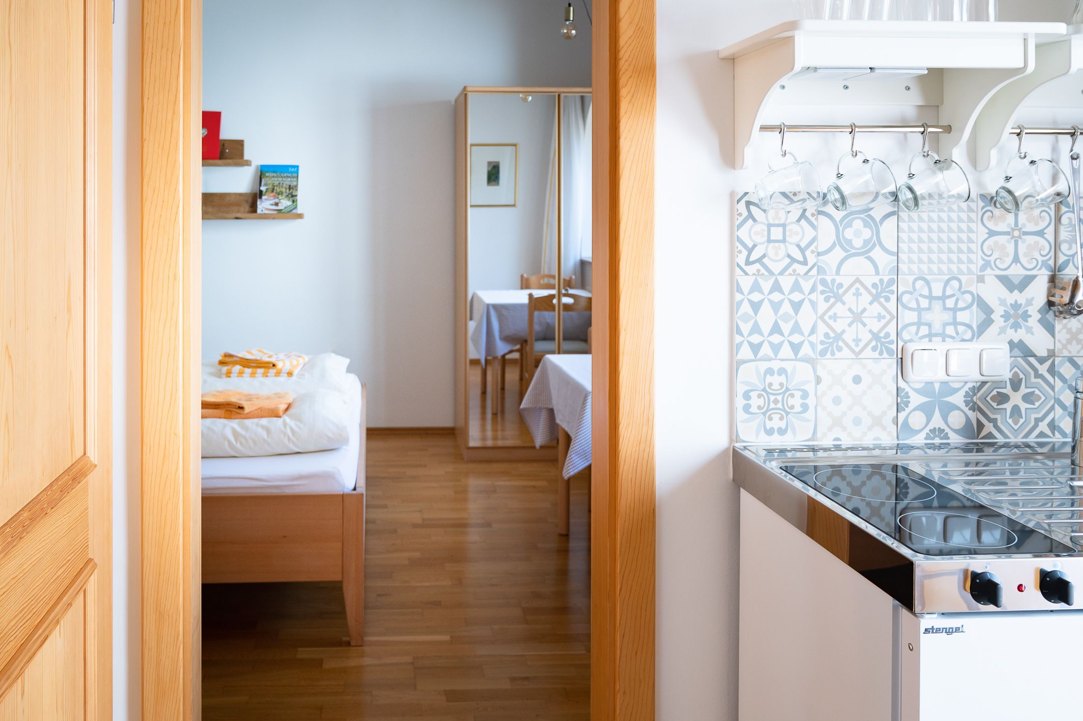 Pohľad do interiéru izby s posteľou, zrkadlom, stolom a malou kuchynkou so vzorovanými dlaždicami.