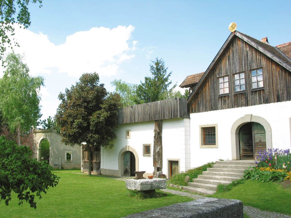 Tradičná budova s drevenou strechou a bielou fasádou, obklopená zelenou záhradou.