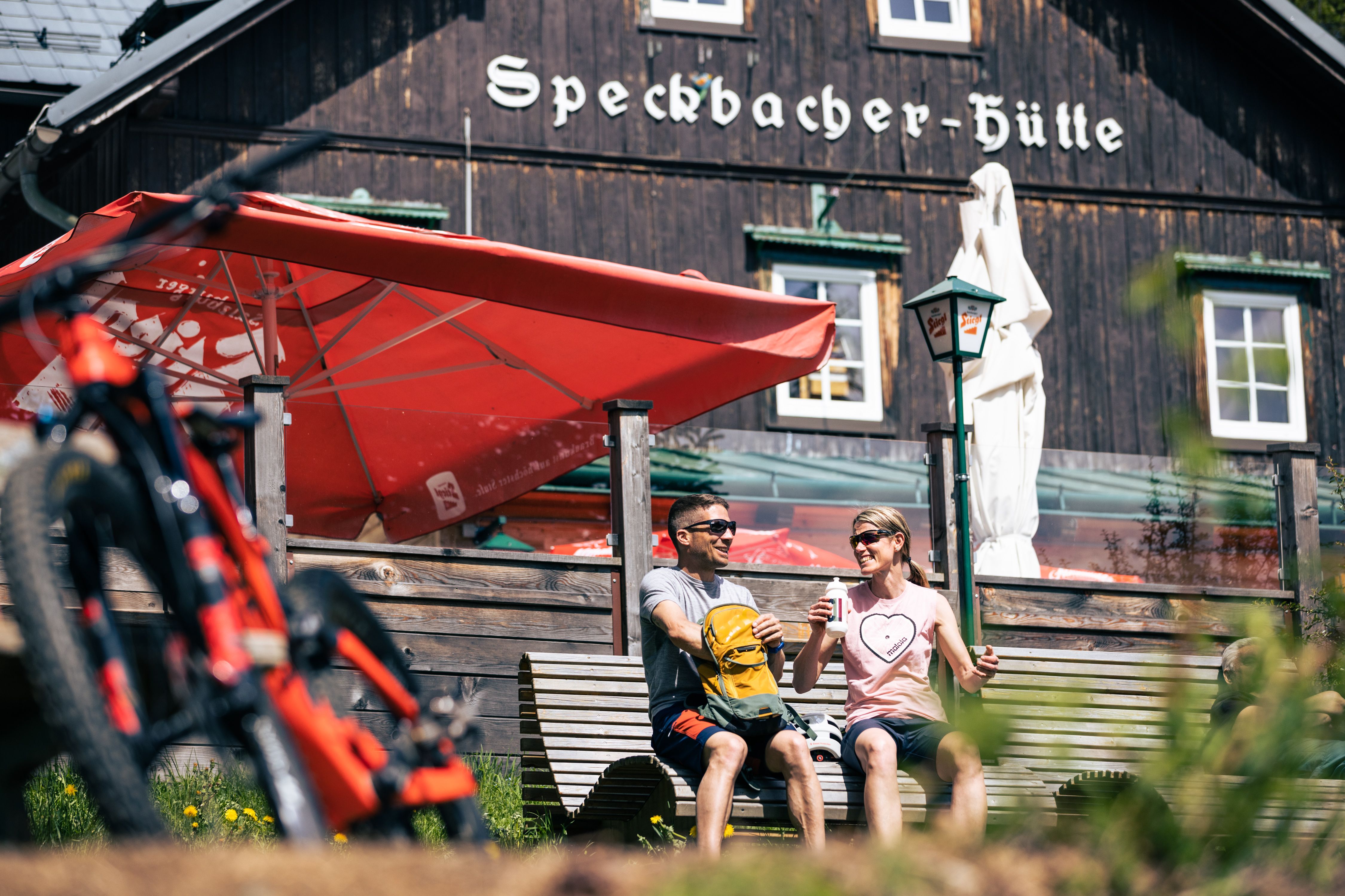 Dvaja ľudia sedia na lavičke pred chatou Speckbacher, v popredí horský bicykel.
