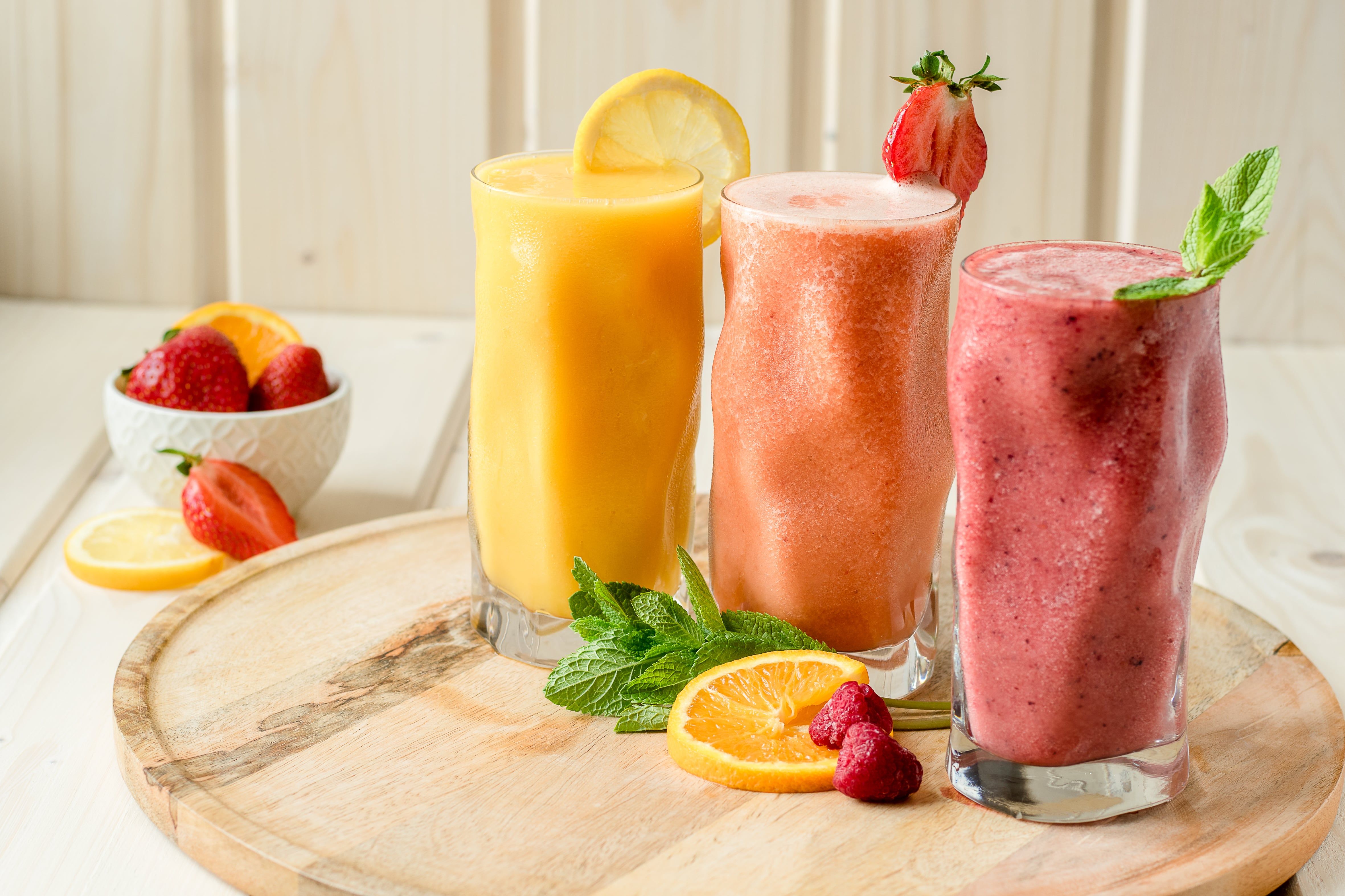 Tri smoothies v pohároch na drevenej doske, zdobené ovocím a mätou.