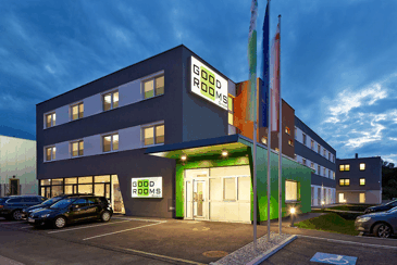 Moderná budova s nápisom "Good Rooms" za súmraku, obklopená autami a vlajkami.