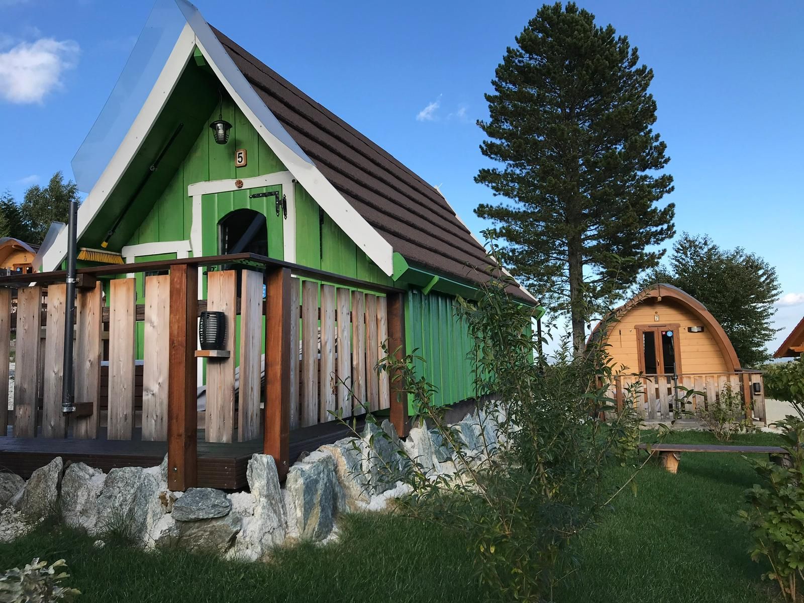 Dve malé drevené chatky v štýle glampingu so zeleno-hnedou fasádou, obklopené stromami a trávou.