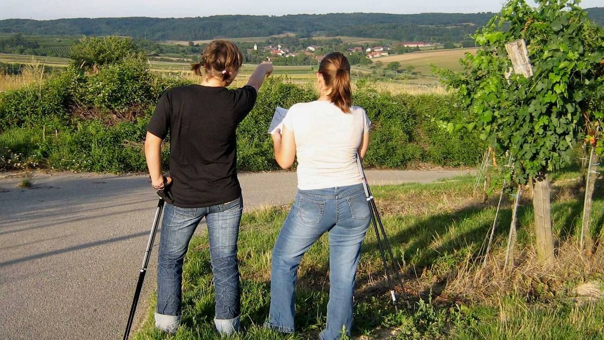 Dve ženy s paličkami stoja na ceste a pozerajú do diaľky, jedna ukazuje smerom k dedine.