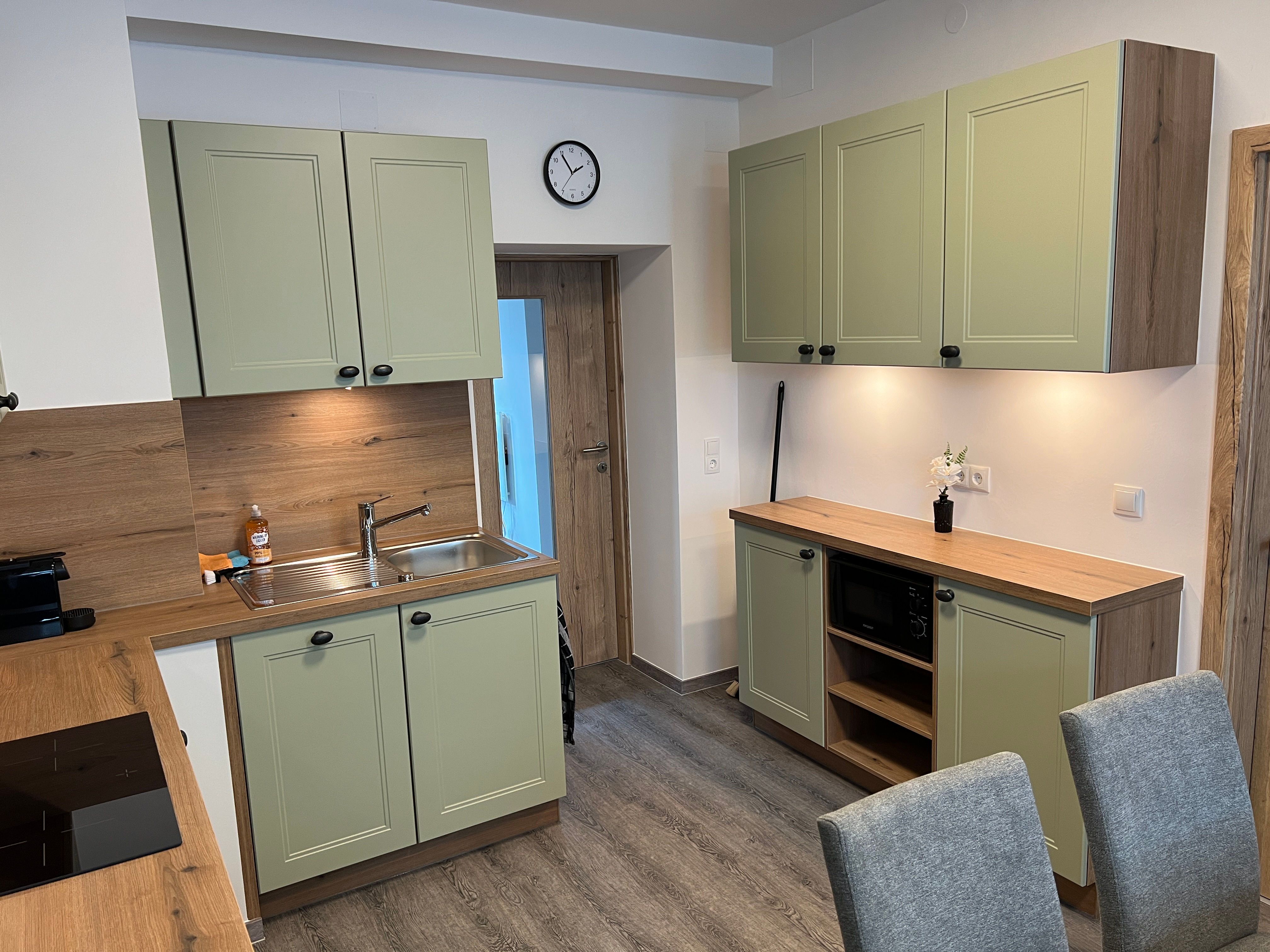 Moderná kuchyňa so zelenými skrinkami, drevenými pracovnými doskami a sivými stoličkami.