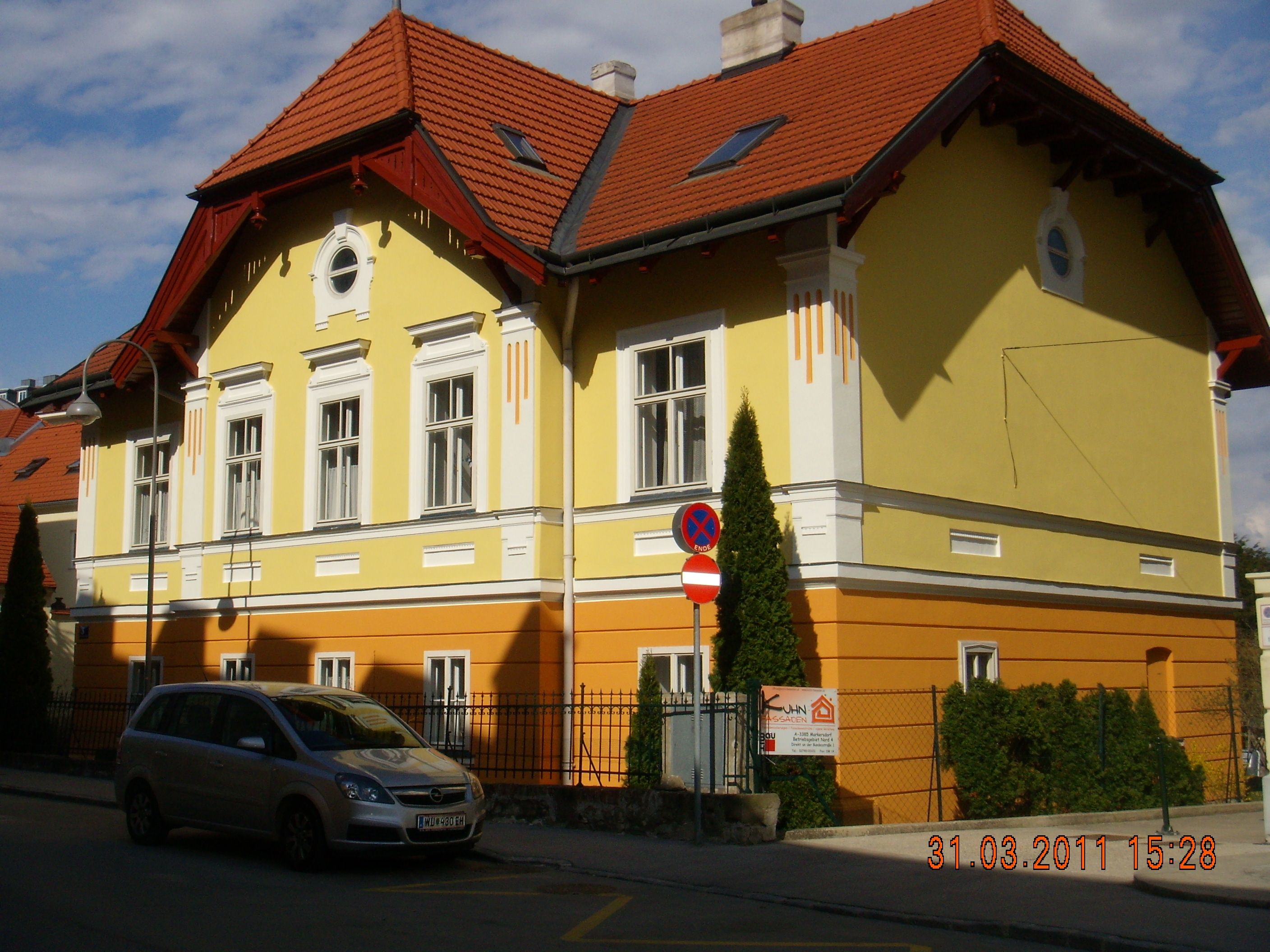 Žltá a oranžová budova s červenou strechou a zaparkovaným autom pred ňou.