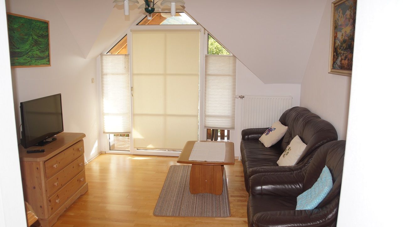 Obývacia izba s koženou pohovkou, dreveným stolom, komodou a TV. Okná so žalúziami, obrazy na stenách.