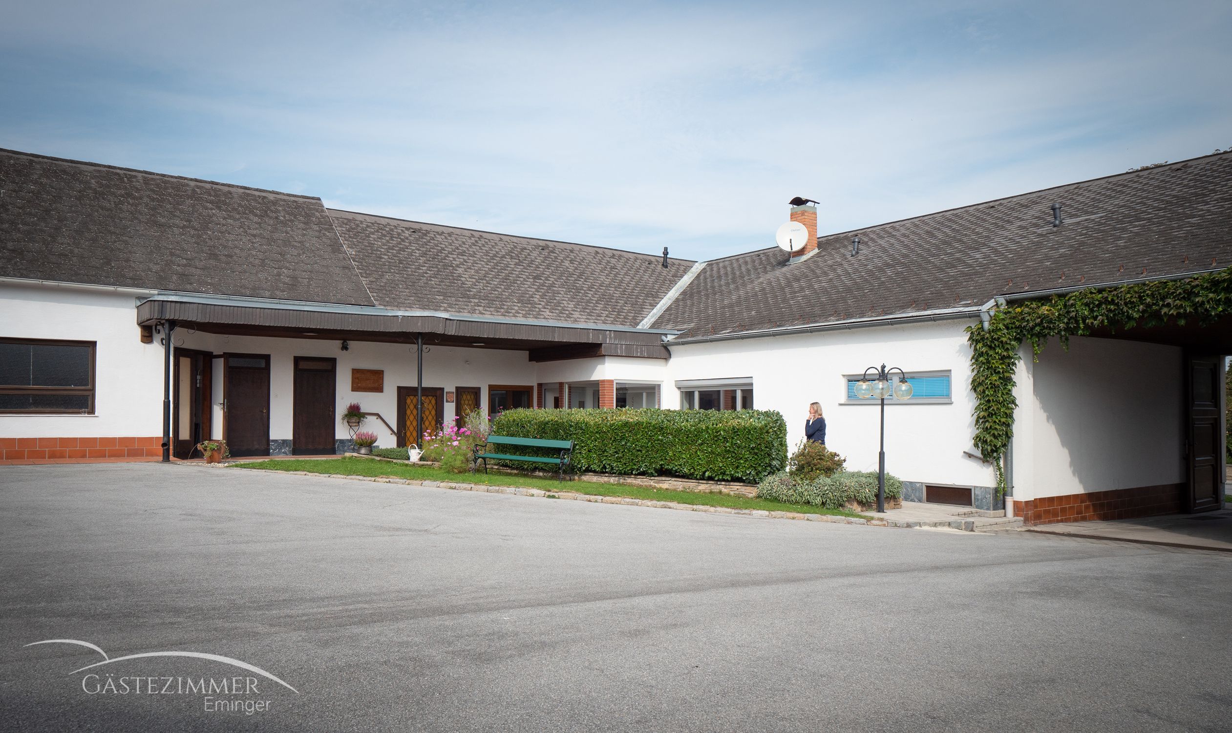 Nádvorie s budovou, parkovacími miestami a živým plotom. Pri živom plote stojí osoba. Na budove je vidieť satelitnú anténu.