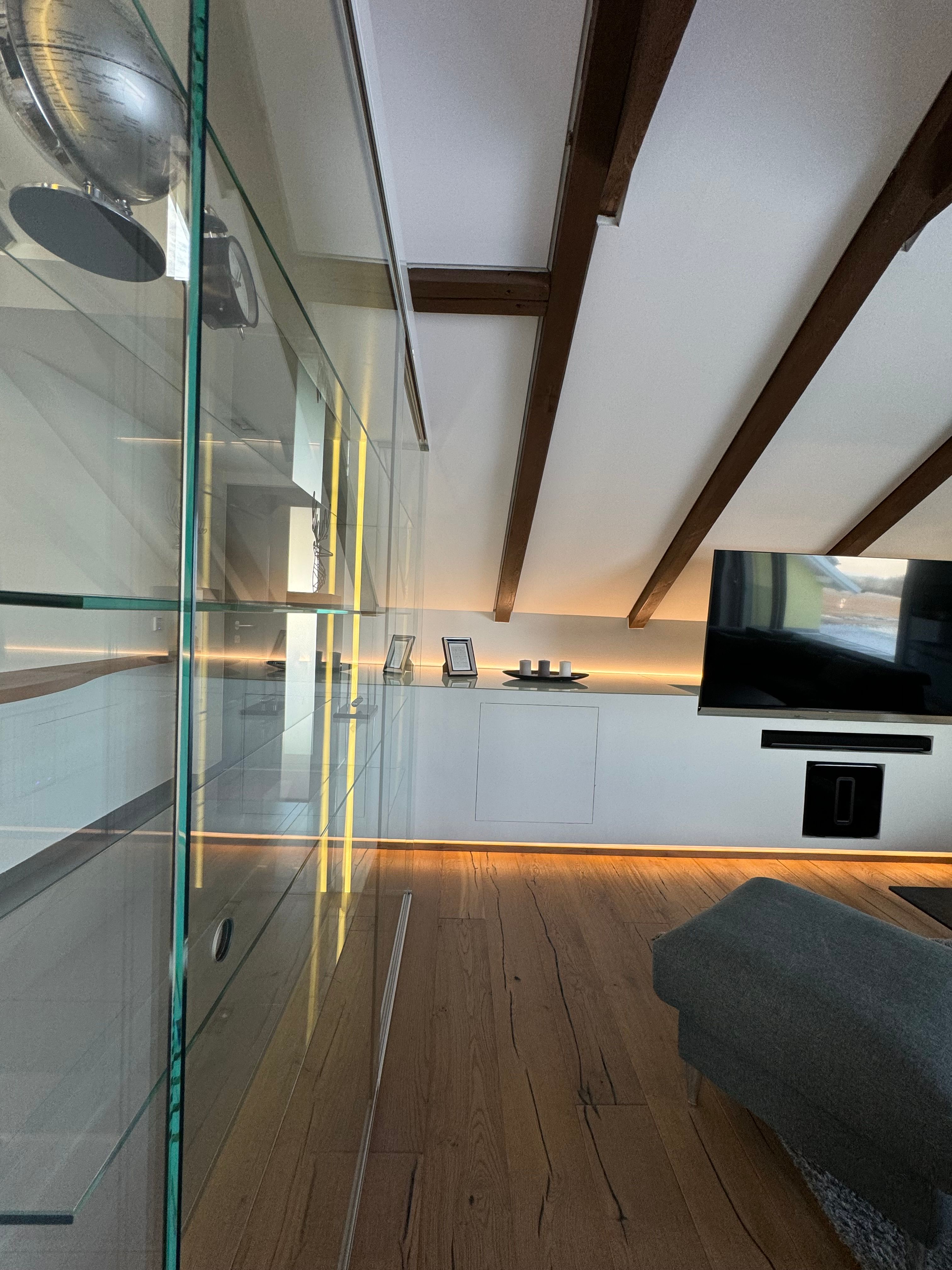 Moderná obývacia izba s drevenou podlahou, presklenou skriňou, TV a šikmým stropom s drevenými trámami.