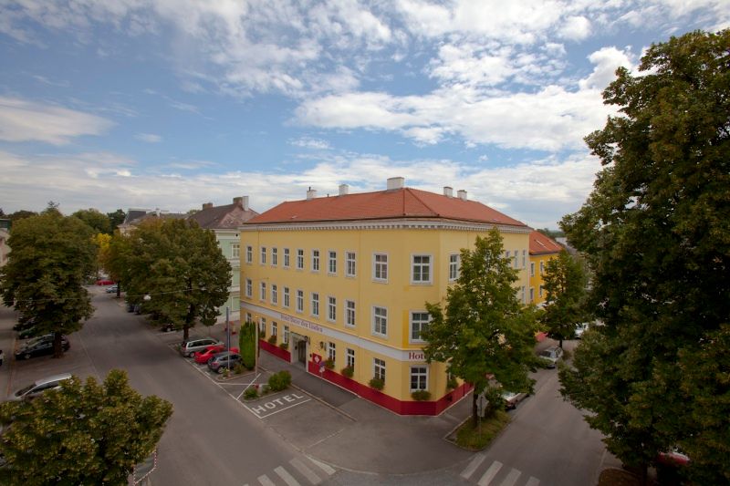 Exteriérový pohľad na žltý hotel s červenou strechou, obklopený stromami a ulicami.