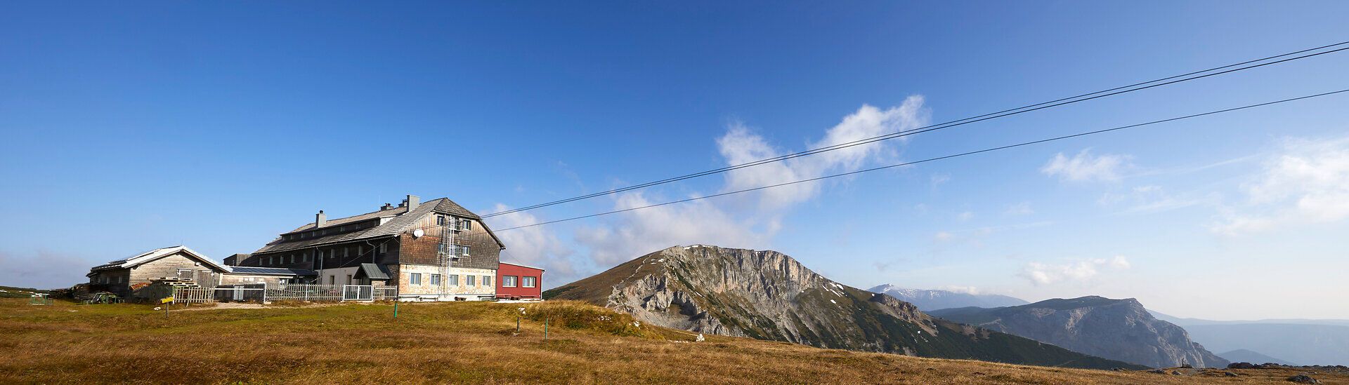 Panoramatický pohľad na horskú chatu s okolitými vrcholmi pod modrou oblohou.