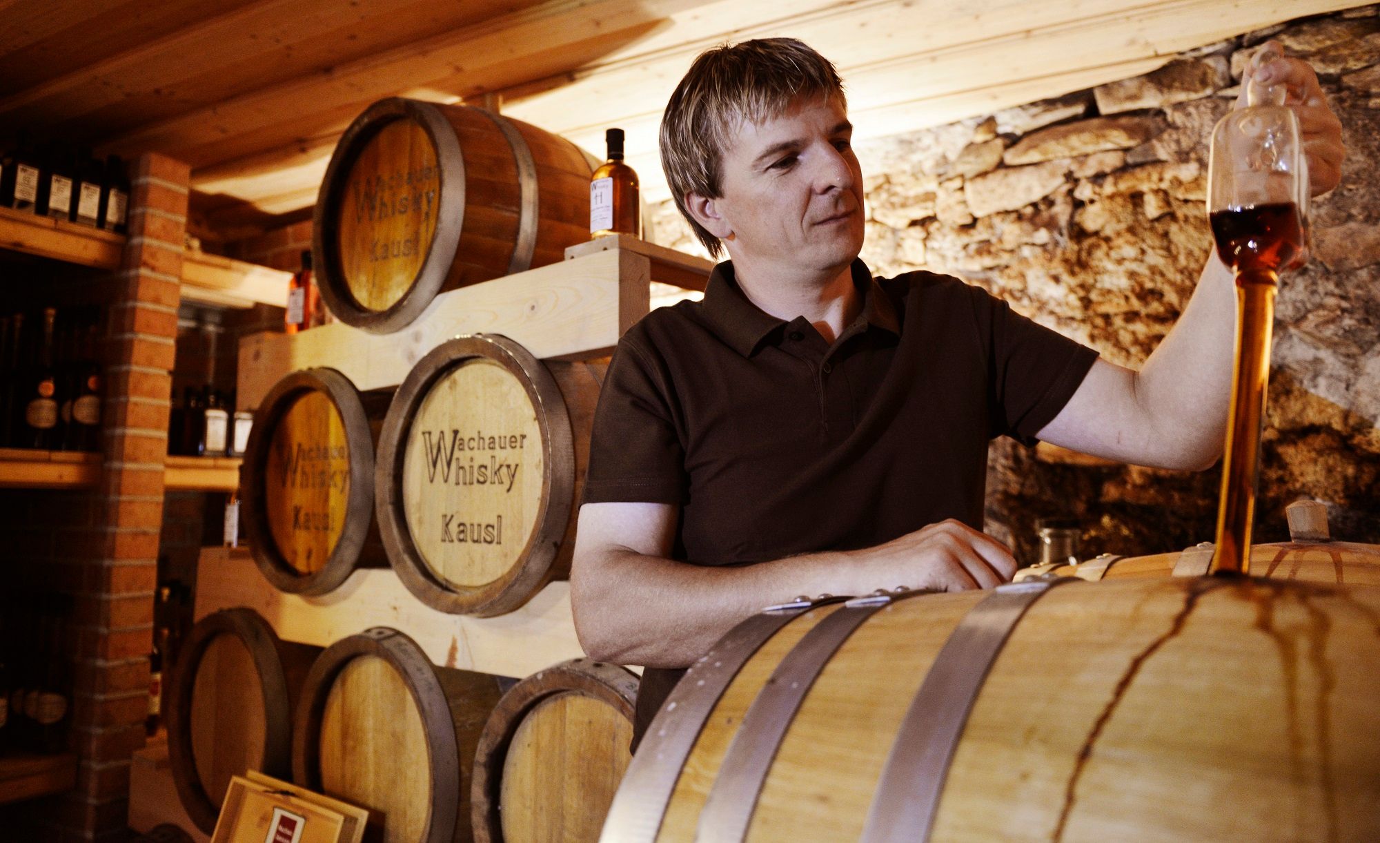 Muž ochutnáva whisky v skladisku so sudmi.