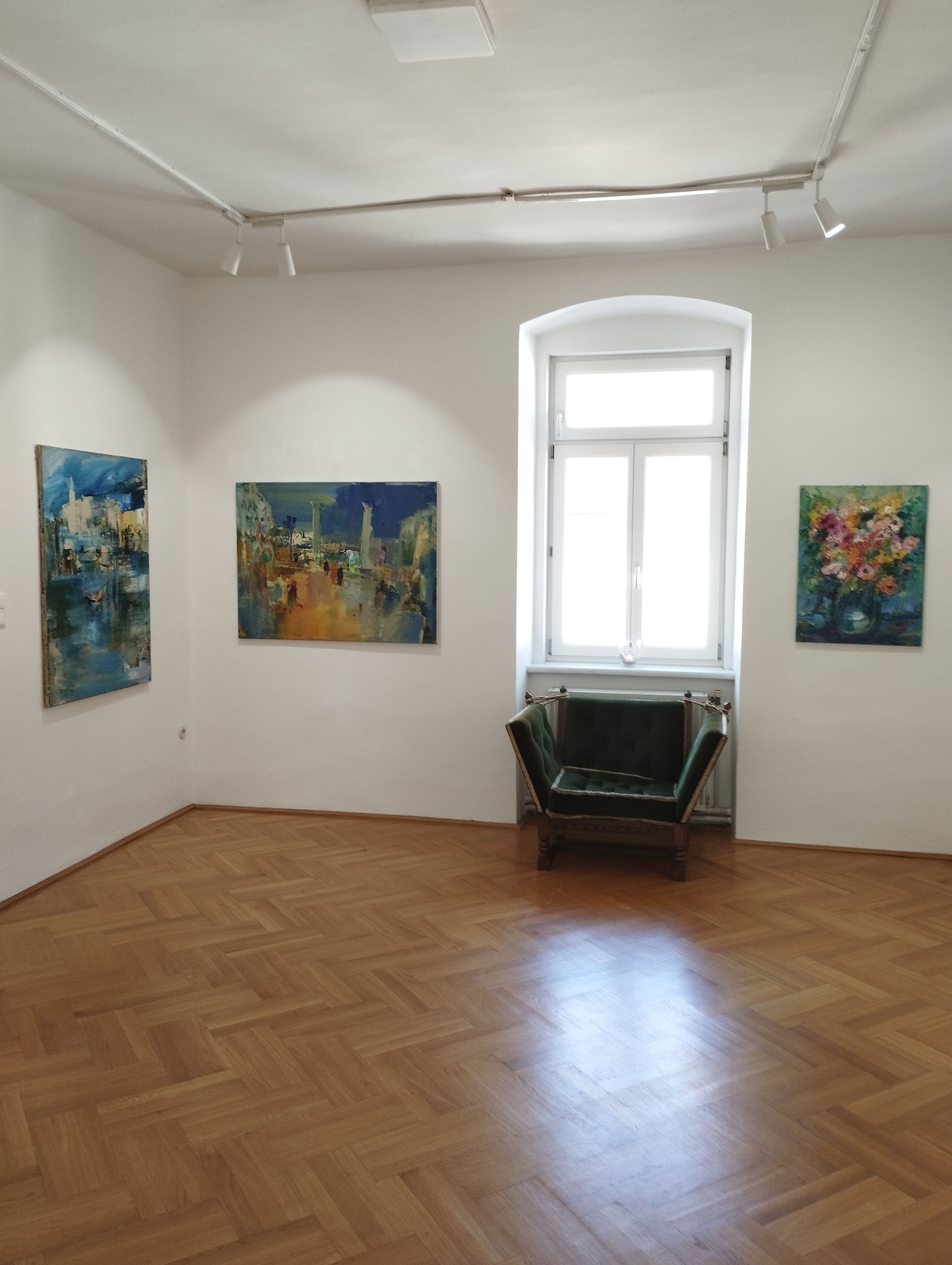 Pohľad do interiéru galérie s tromi obrazmi na stenách a zeleným kreslom pred oknom.