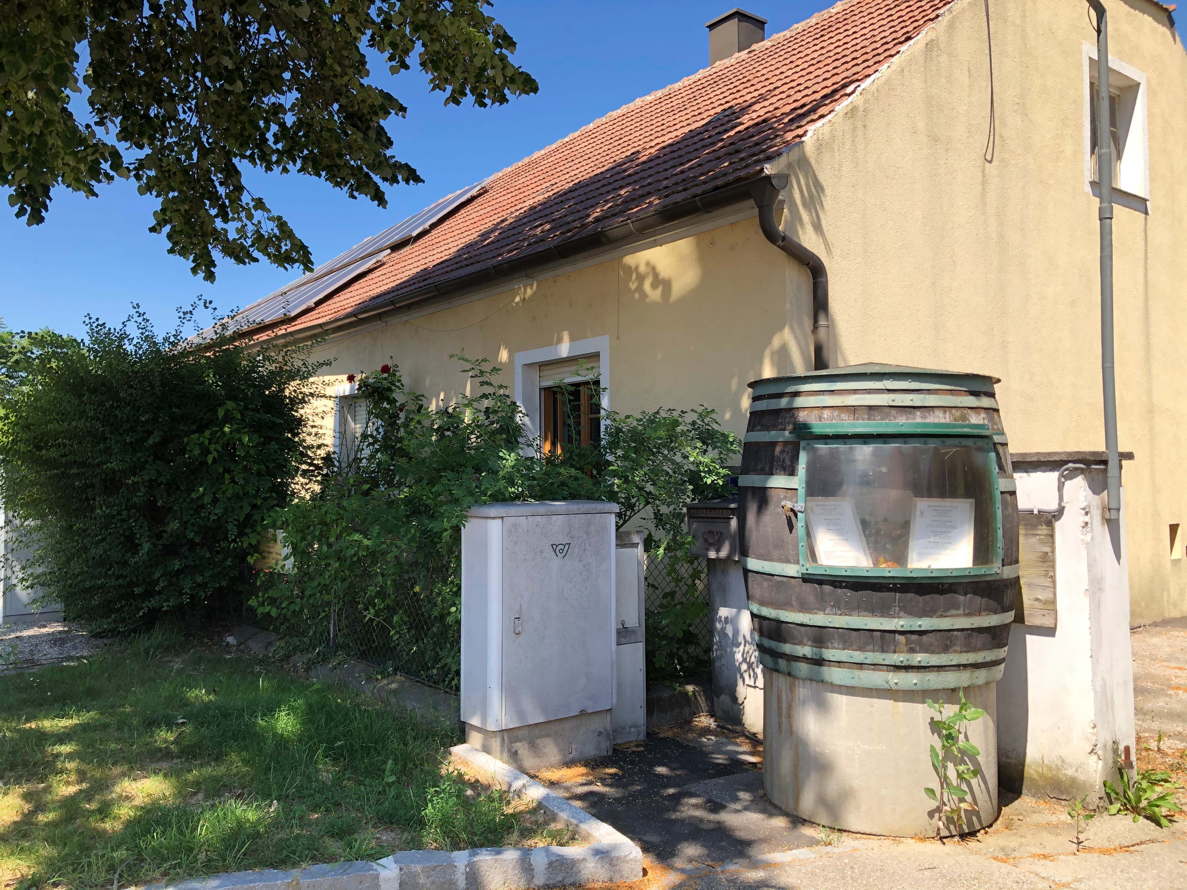 Žltý dom s červenou škridlovou strechou a solárnymi panelmi. V popredí veľký sud s priezorníkom a rastlinami.