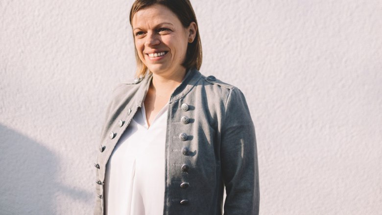 Žena v sivej bunde pred bielou stenou.
