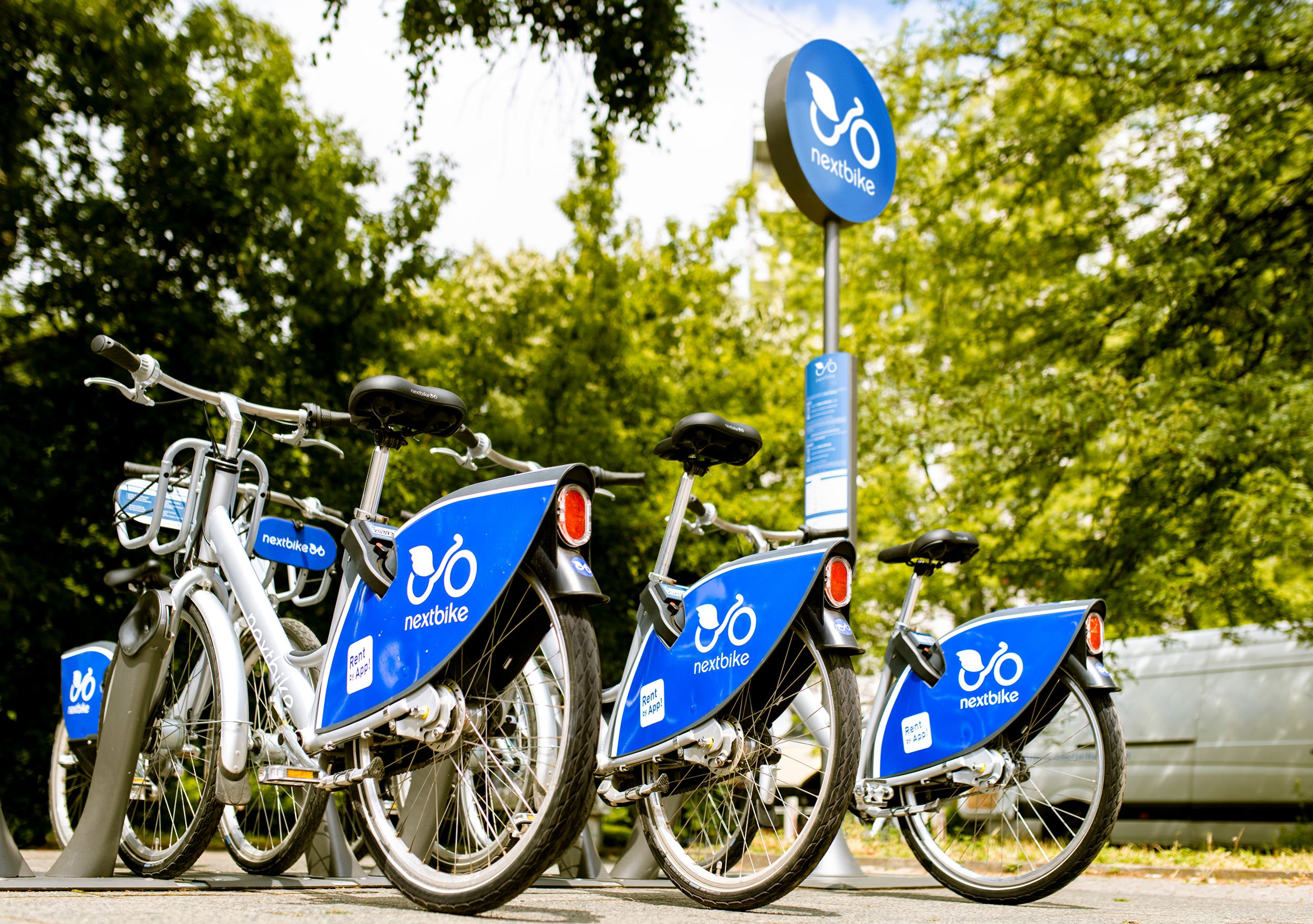 Niekoľko bicyklov Nextbike na vonkajšej stanici obklopenej stromami.