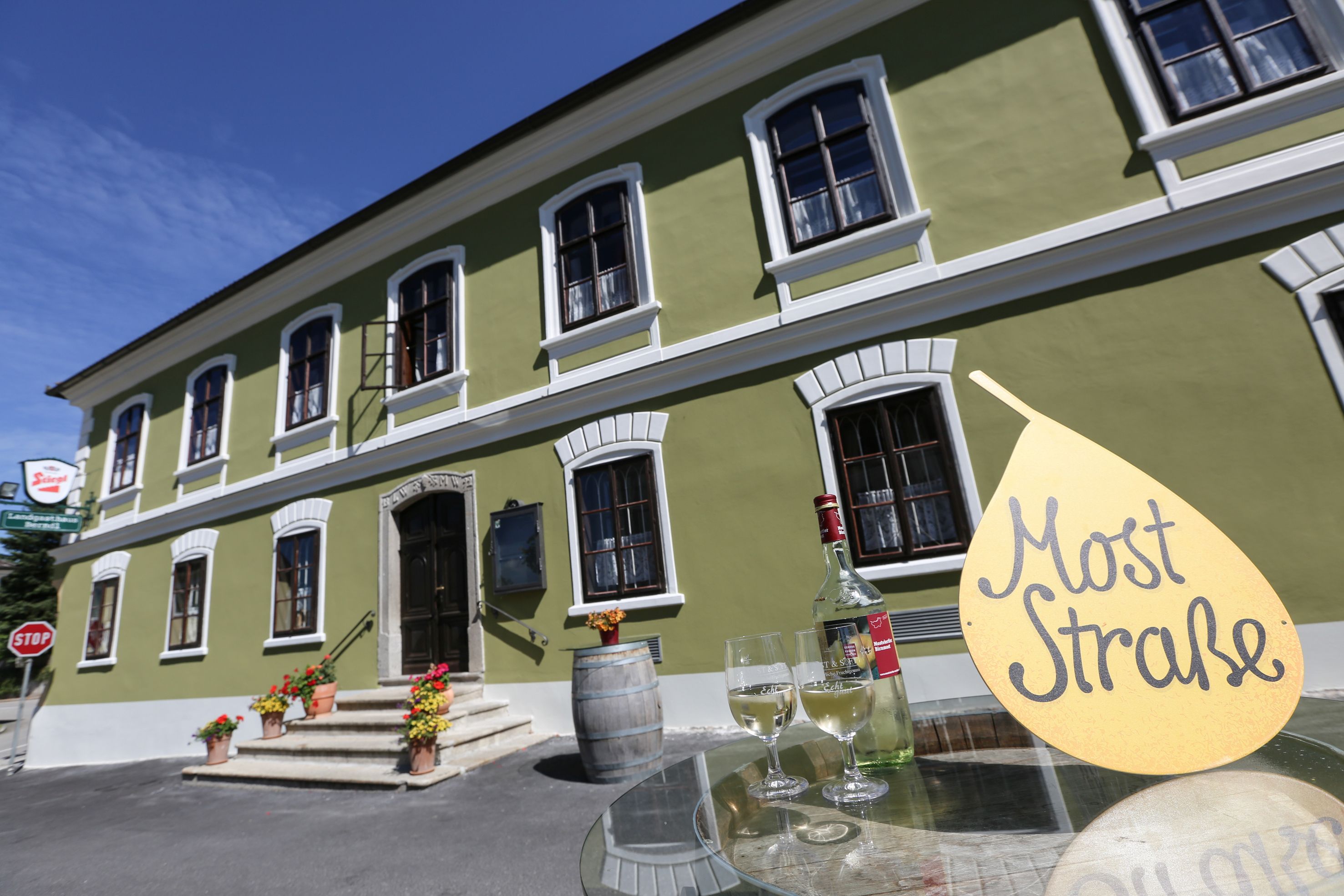 Zelená budova s bielymi oknami, pred ňou stôl s fľašou vína a pohármi, tabuľka s nápisom "Mušt Straße".