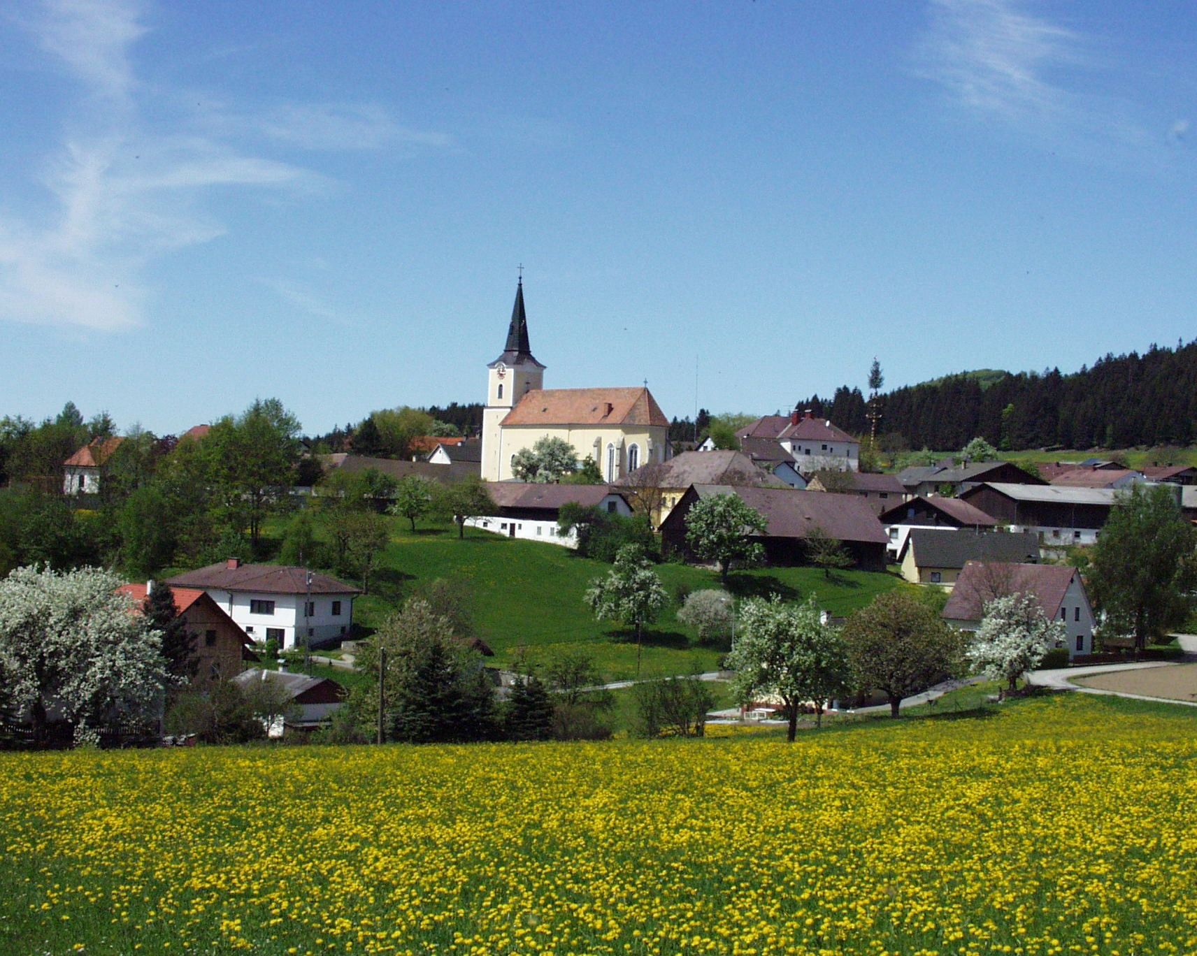 Krajina s kostolom a dedinou v pozadí, obklopená kvitnúcimi stromami a lúkami.