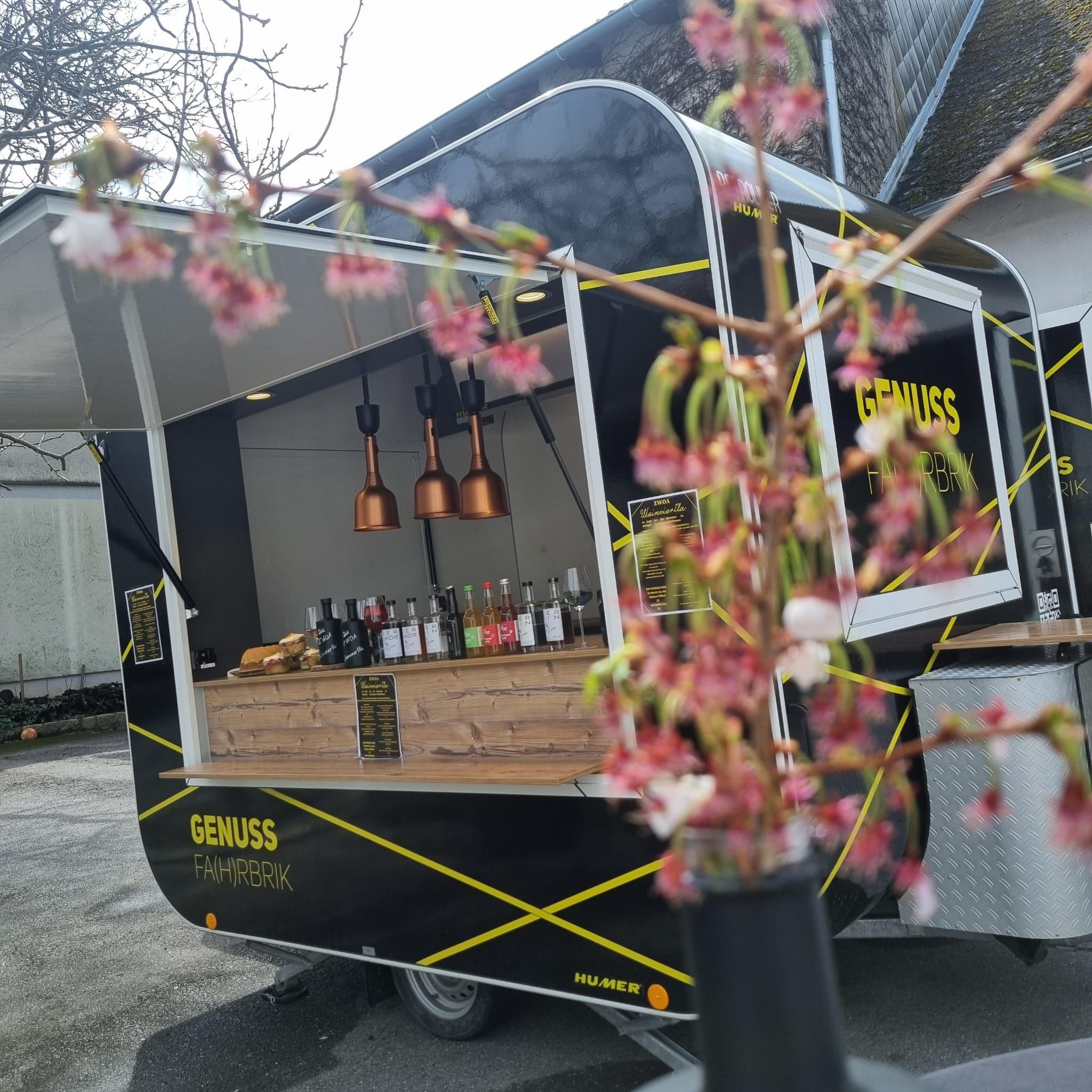 Vonku parkuje čierne nákladné auto s nápisom "Genuss Fahrbrik". V popredí vidno rozmazané kvety.
