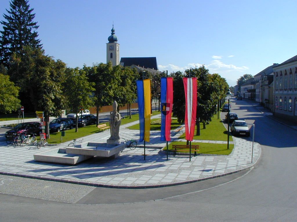 Námestie v Ruprechtshofene s kostolom, sochami a vlajkami.