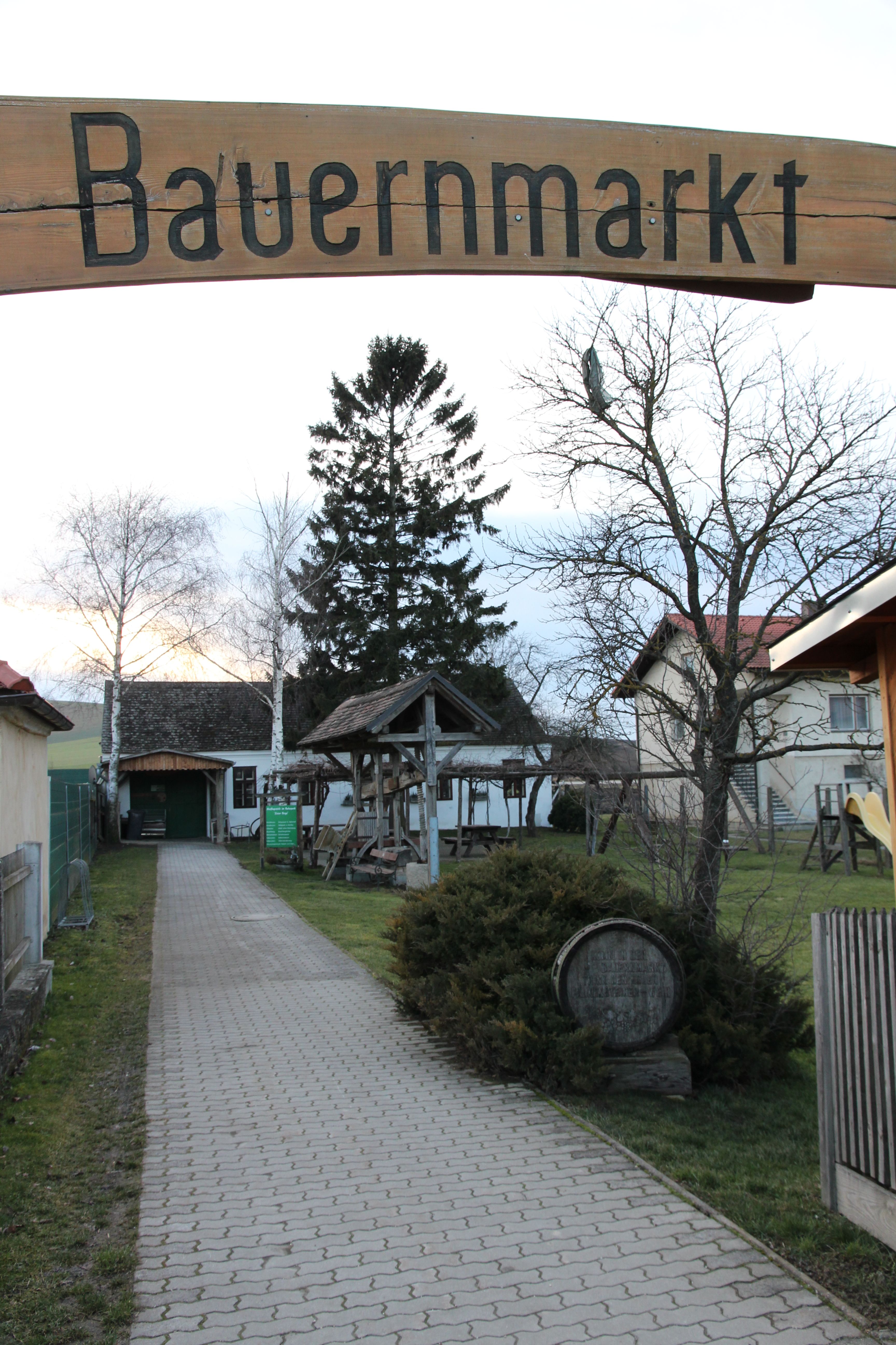 Vchod na farmársky trh s drevenou tabuľou a vydláždenou cestou obklopenou stromami a budovami.