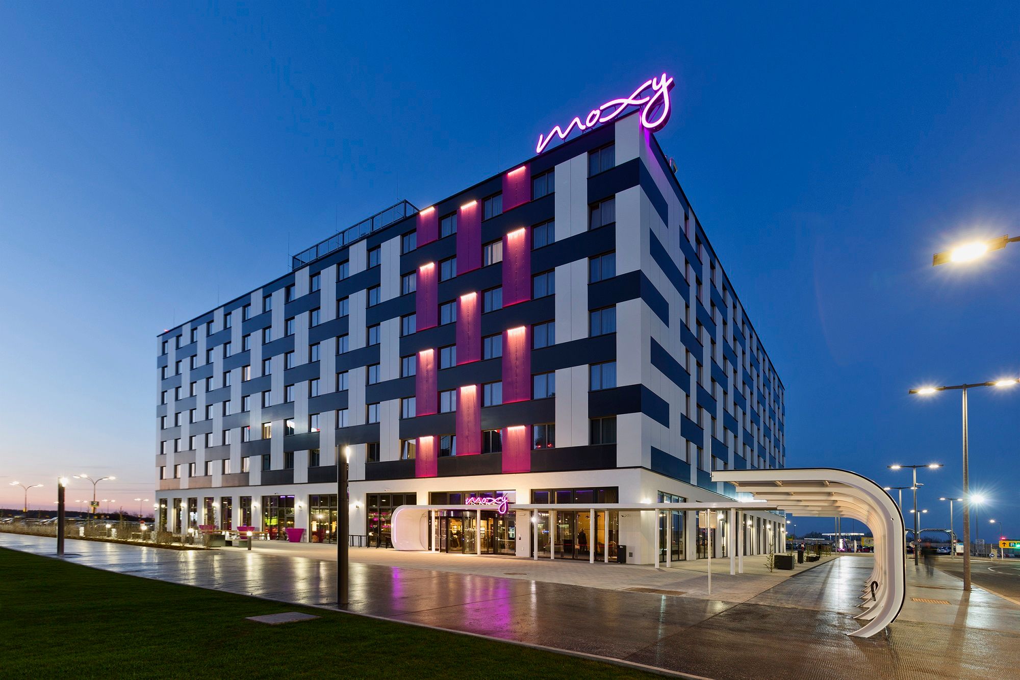 Pohľad na exteriér hotela Moxy Vienna Airport za súmraku s osvetlenými nápismi.