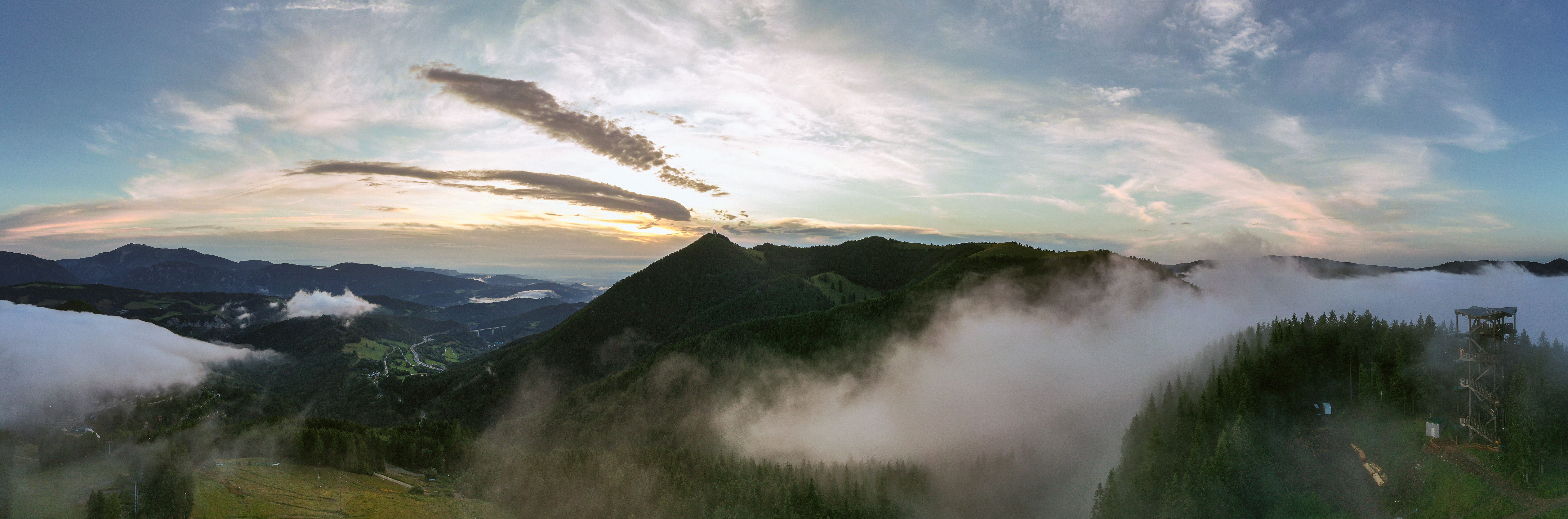Panoramatický pohľad na Sonnwendstein pri východe slnka s hmlou a rozhľadňou v popredí.