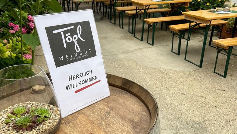 Na sude s vínom je nápis "Weingut Tögl" a "Herzlich Willkommen" ("Vitajte"). V pozadí je vidieť pivné stoly a rastliny.