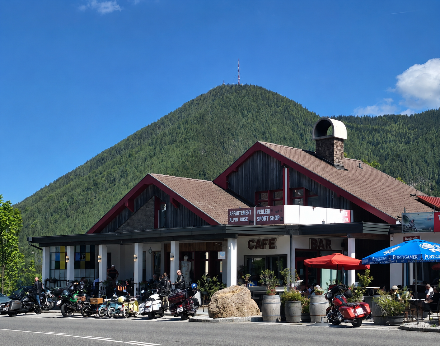 Kaviareň a bar s motorkami pred domom, obklopené horami a stromami.