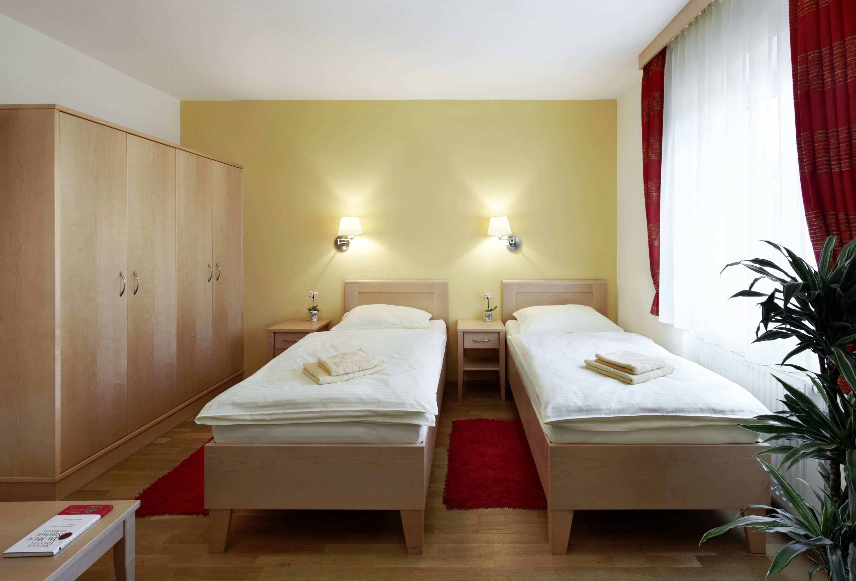 Izba pre hostí s dvoma samostatnými posteľami, veľkou skriňou, červenými závesmi a rastlinou.