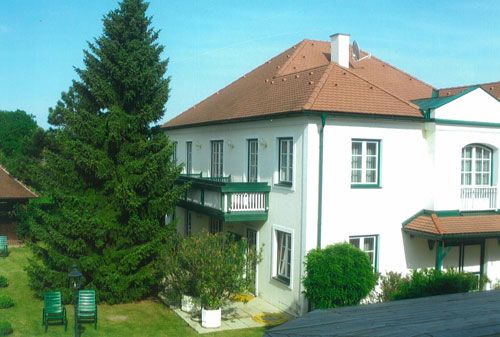 Dvojpodlažný biely penzión s červenou strechou a zelenými okenicami, obklopený záhradou so stromami a záhradným nábytkom.
