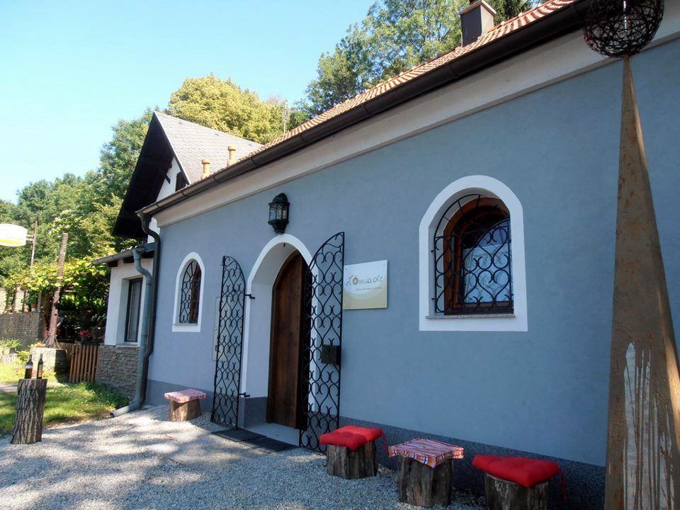 Malá modrá budova s drevenými dverami a ozdobnými mrežami pred oknami. Pred ňou stoja drevené stoličky s červenými vankúšmi.