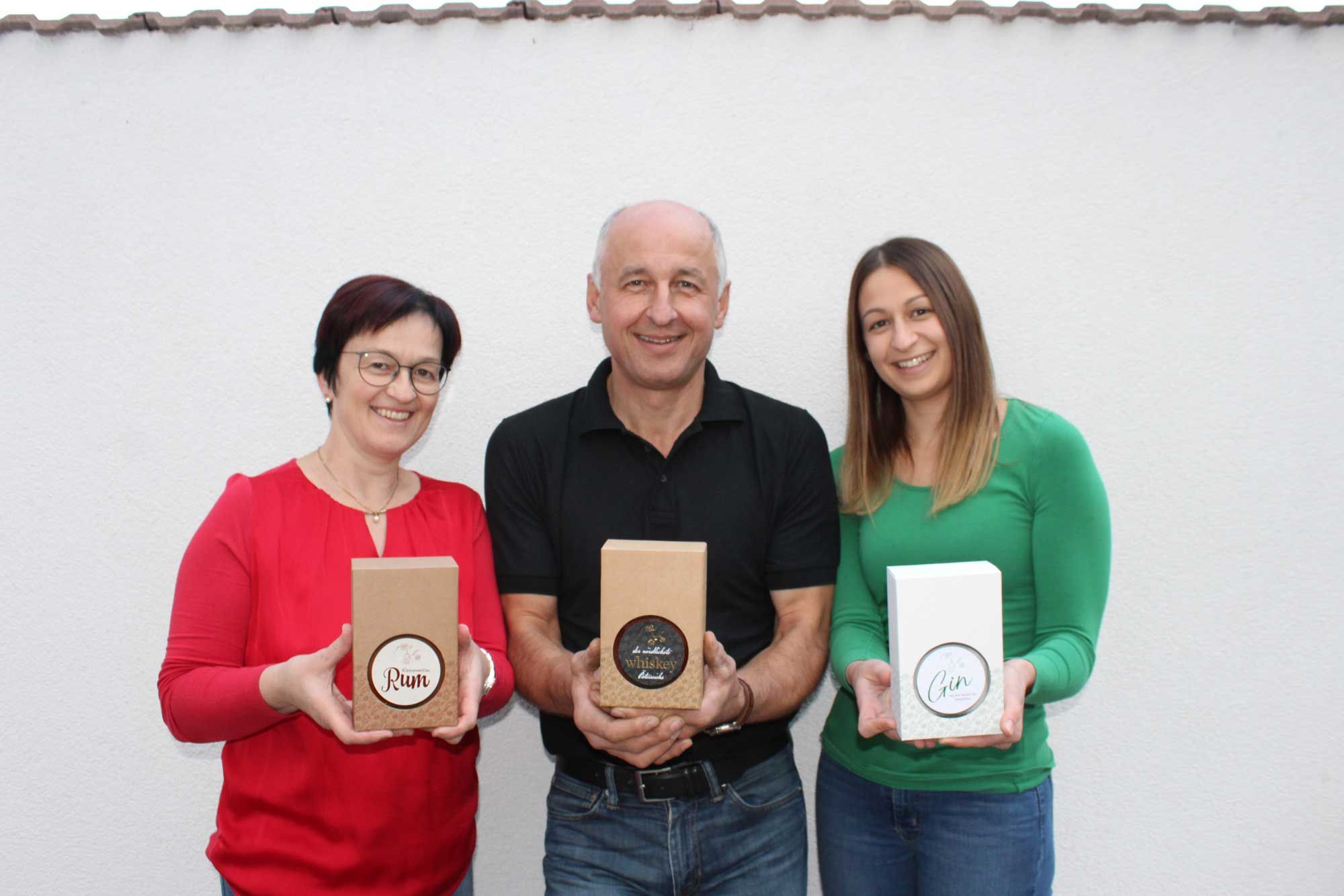 Tri osoby držia pred bielou stenou darčekové škatule s rumom, whisky a ginom.