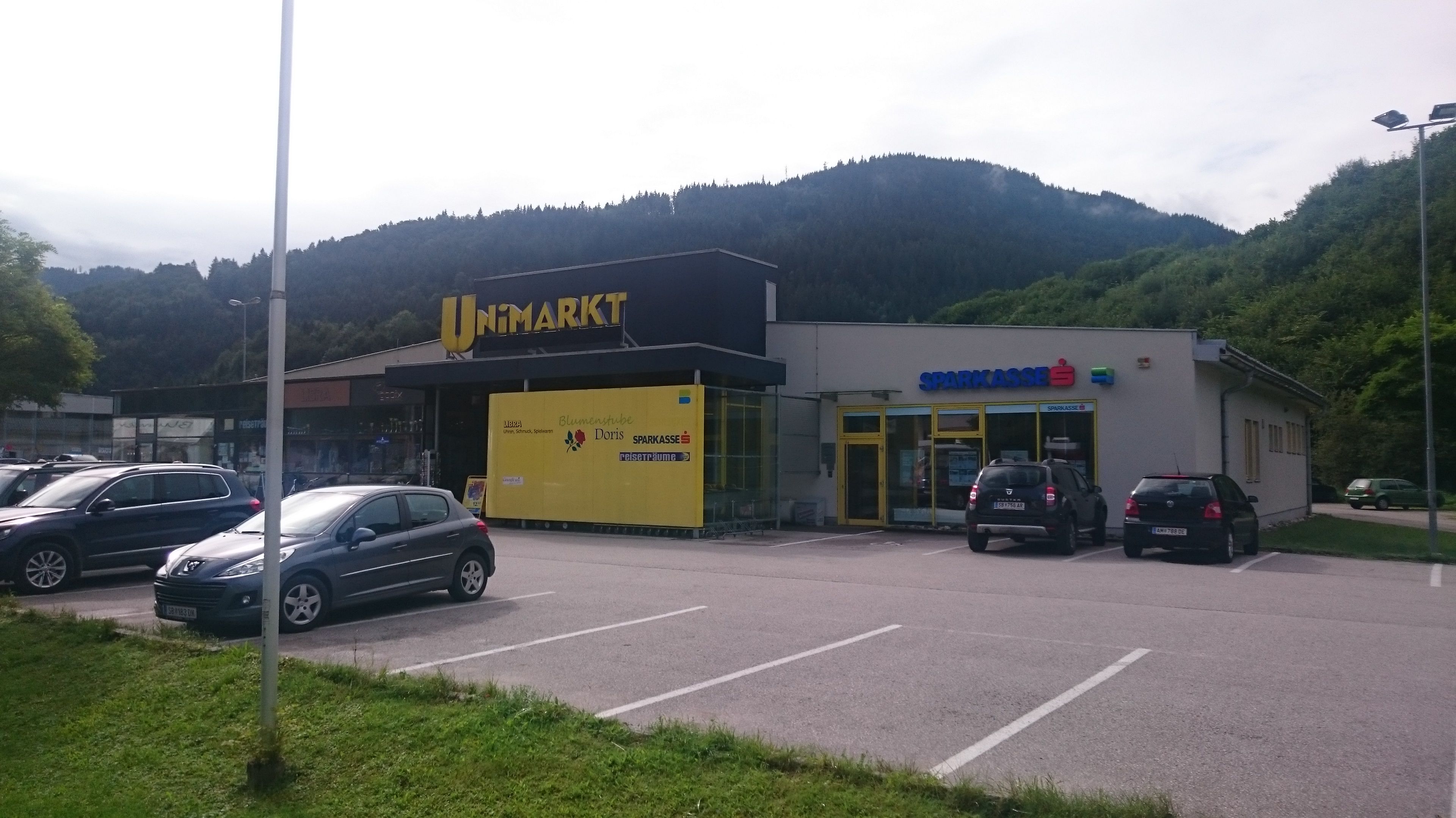 Nákupné centrum so supermarketom Unimarkt a sporiteľňou pred zalesnenými kopcami.