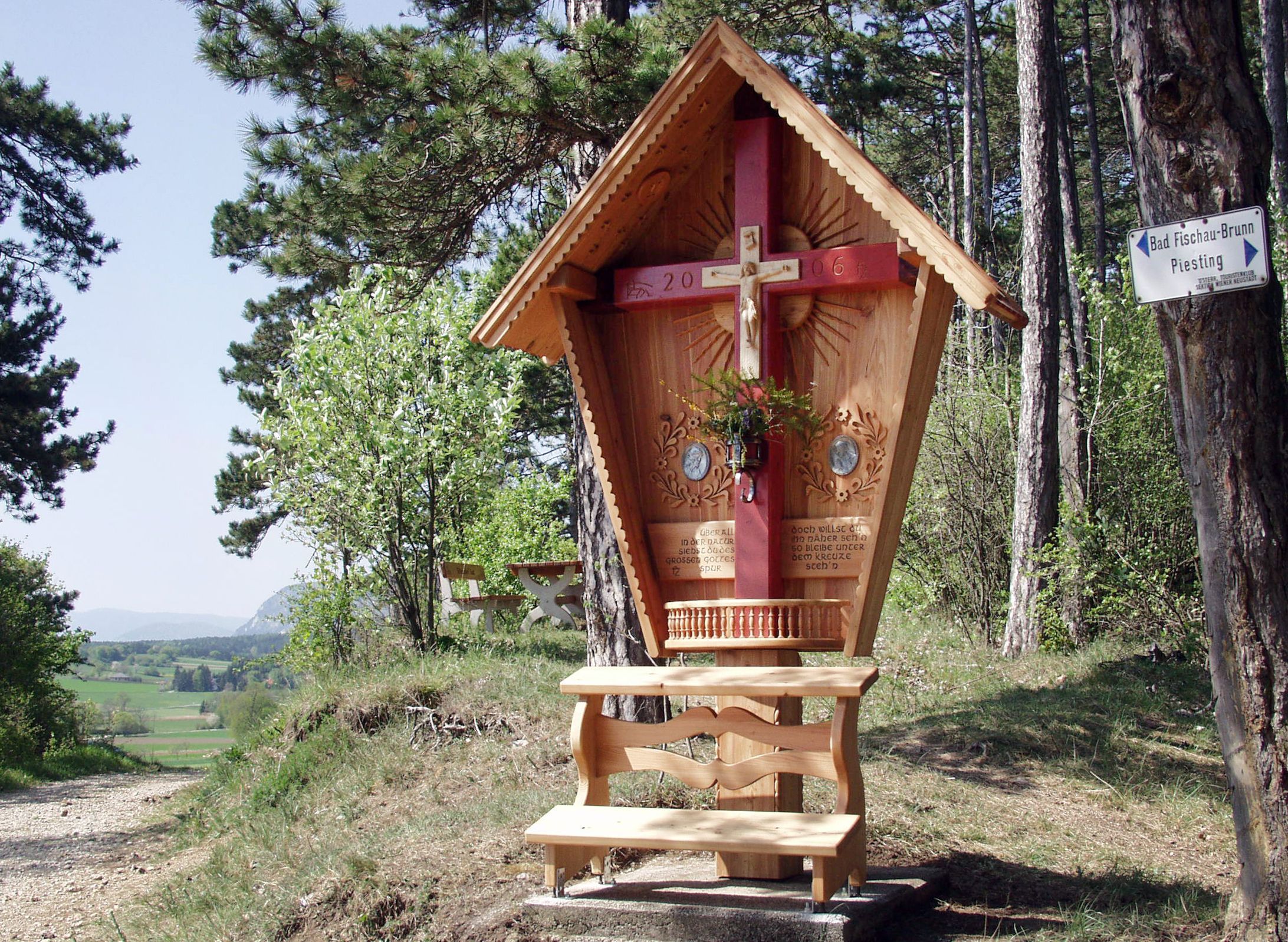 Drevený kríž s červeným trámom a vyrezávanou strechou v lesnej krajine.