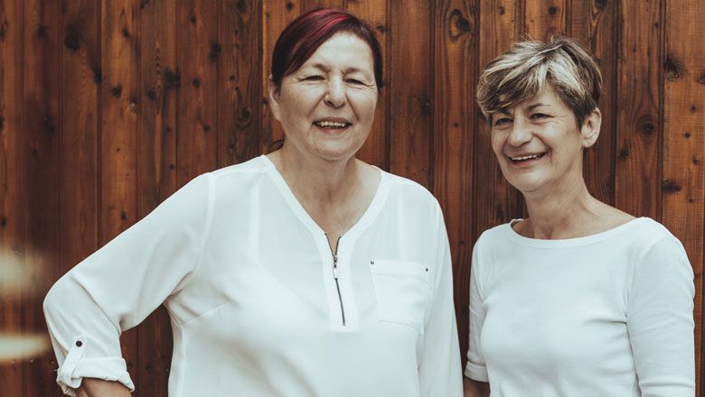 Dve ženy v bielych blúzkach stoja pred drevenou stenou a usmievajú sa.