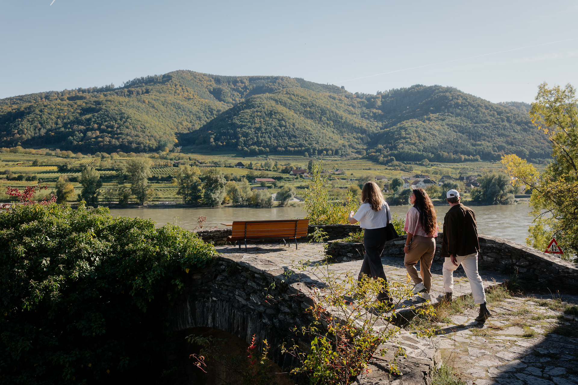 Drei Personen gehen über einen steinernen Weg an einem Aussichtspunkt; vor ihnen liegt die Donau mit grünen Hügeln und Feldern im warmen Tageslicht.
