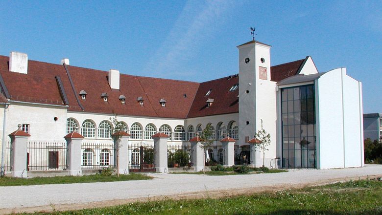 Zámok Katzelsdorf s modernou prístavbou a historickou budovou. Biela fasáda so zaoblenými oknami, červená strecha a veľké vnútorné nádvorie