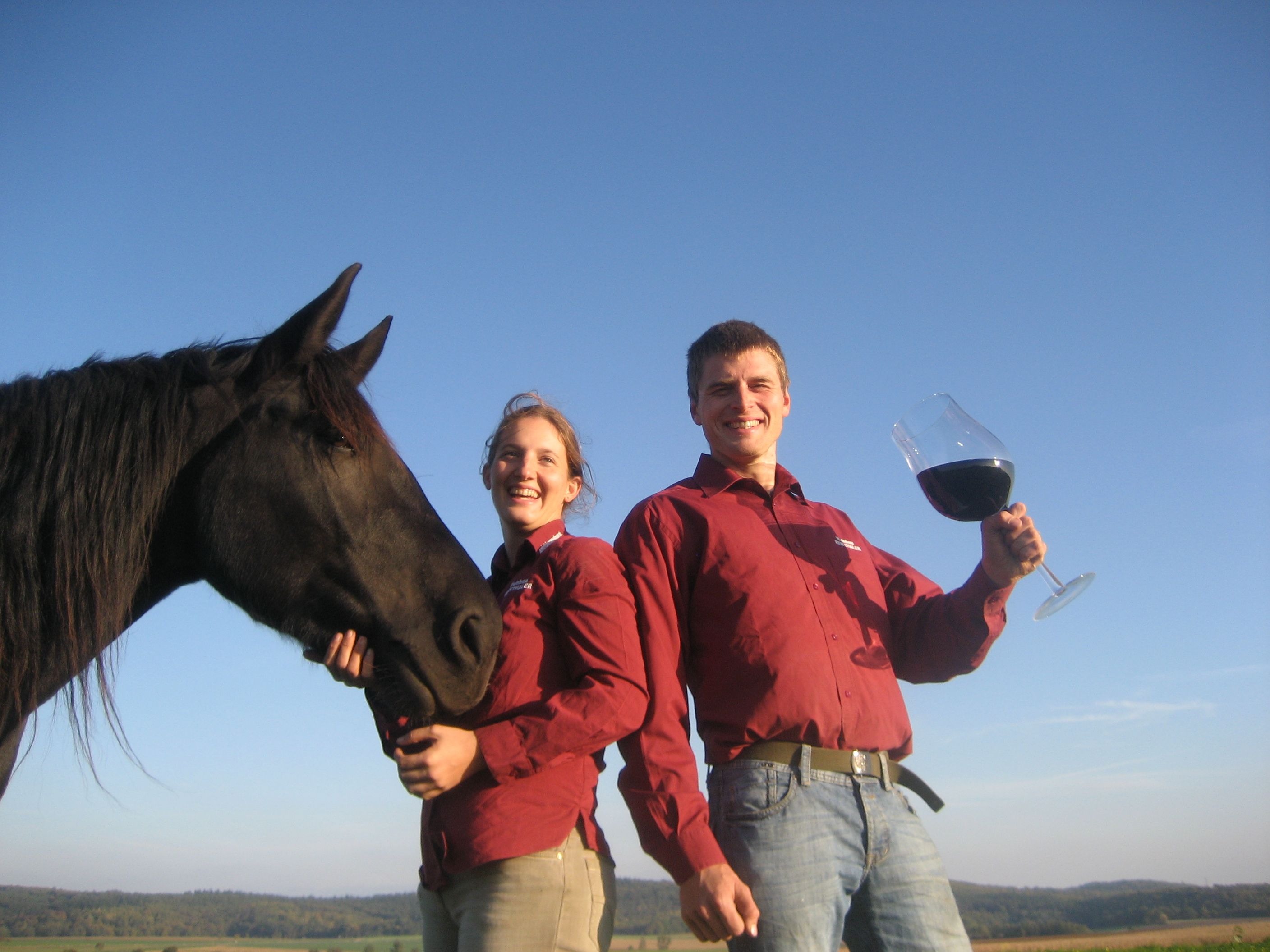 Dvaja ľudia v červených košeliach s čiernym koňom a veľkým pohárom na víno vonku.