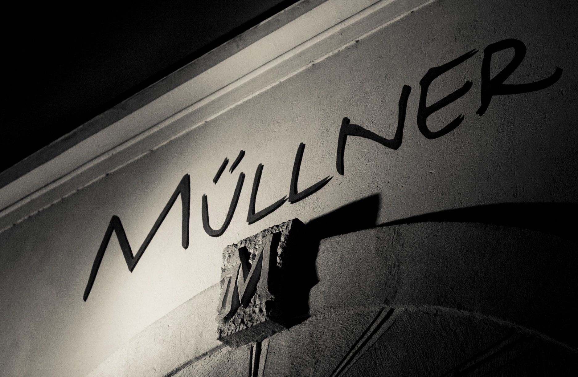 Čiernobiela fotografia nápisu "Müllner" na stene s ozdobným písmenom "M".