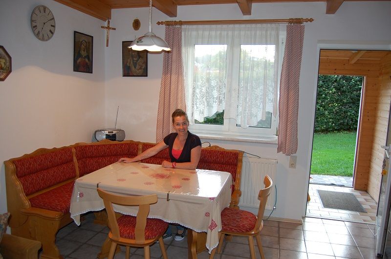 Žena sedí pri jedálenskom stole v útulnej kuchyni s rohovou lavicou a červenými vankúšmi.