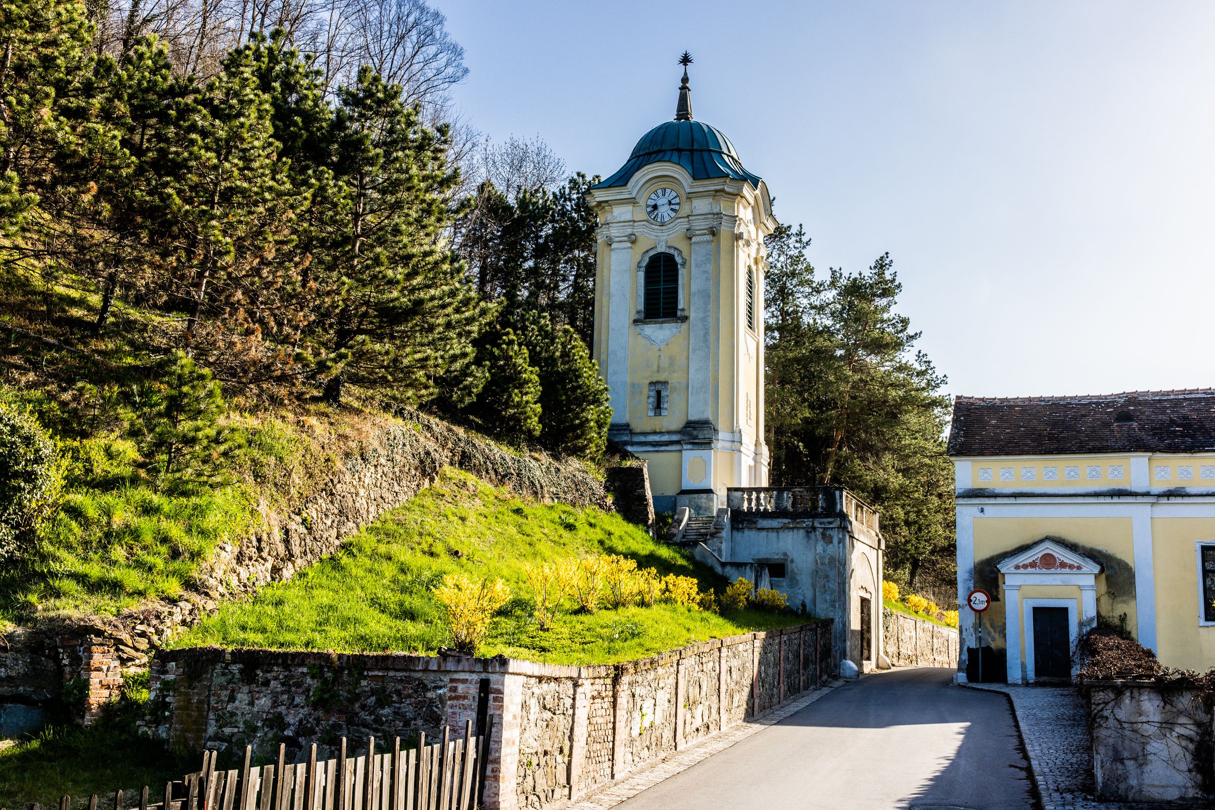 Žltá veža s hodinami v Bad Erlachu, obklopená stromami a slnečnou stráňou, okolo ktorej vedie úzka cesta.