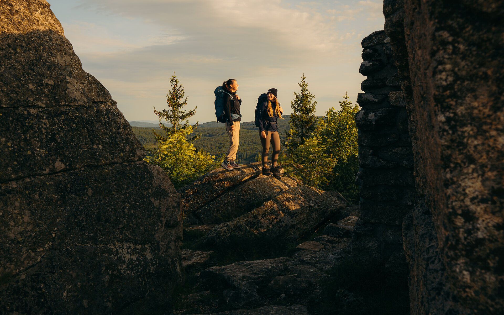 Dvaja turisti stoja medzi impozantnými skalnými útvarmi a v zlatistom rannom svetle sa kochajú výhľadom na lesnú krajinu vo Waldvierteli.
