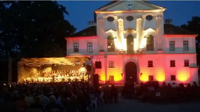 Osvetlená budova s orchestrom hrajúcim na vonkajšom pódiu, obklopená sediacim publikom v noci.